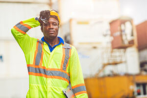 A man in construction gear stands at a worksite and wipes sweat from his face.