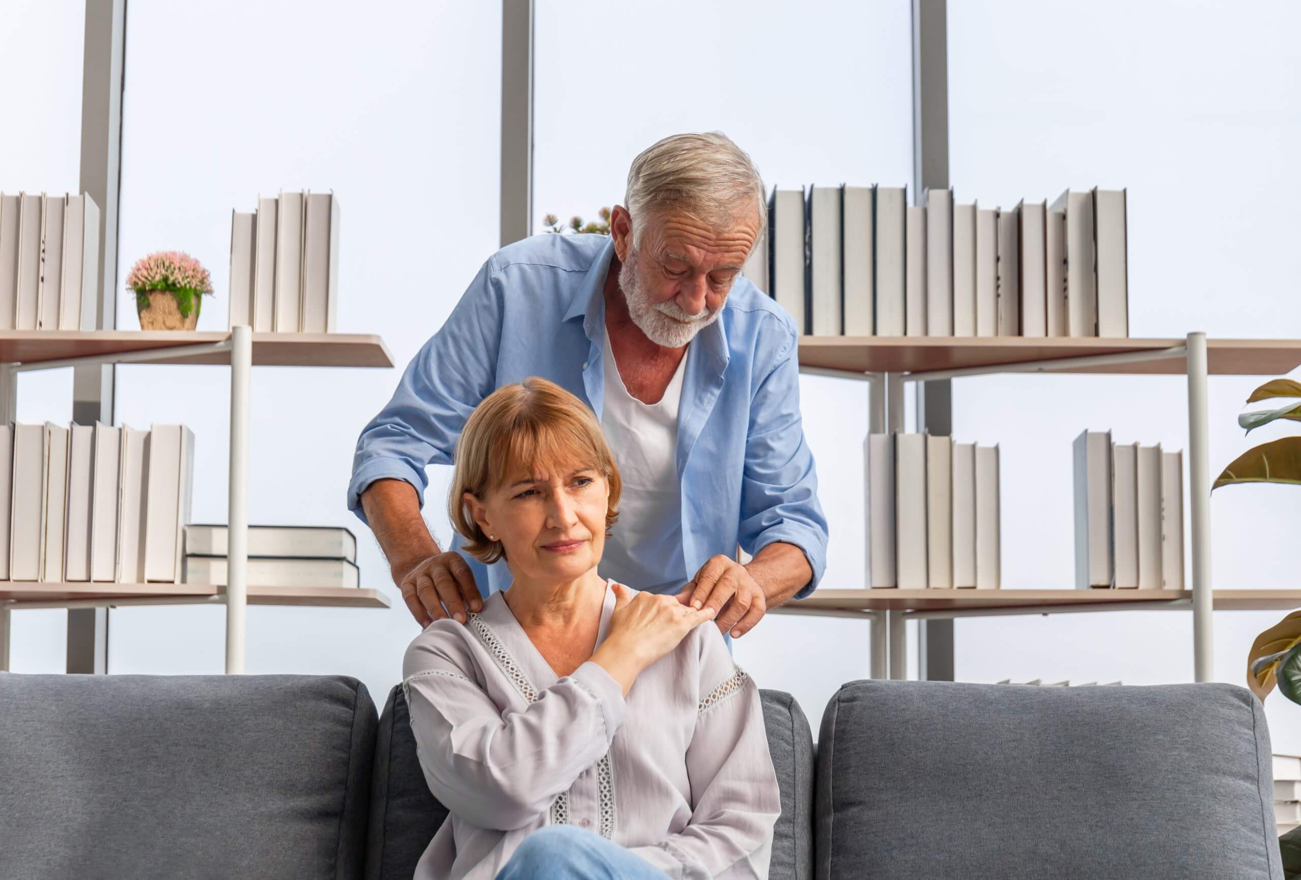An older woman sits on a couch and holds her shoulder in pain while a man leans over her and touches her shoulder. 