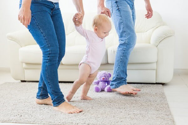 Barefoot parents hold their baby's hands while she takes her first steps.
