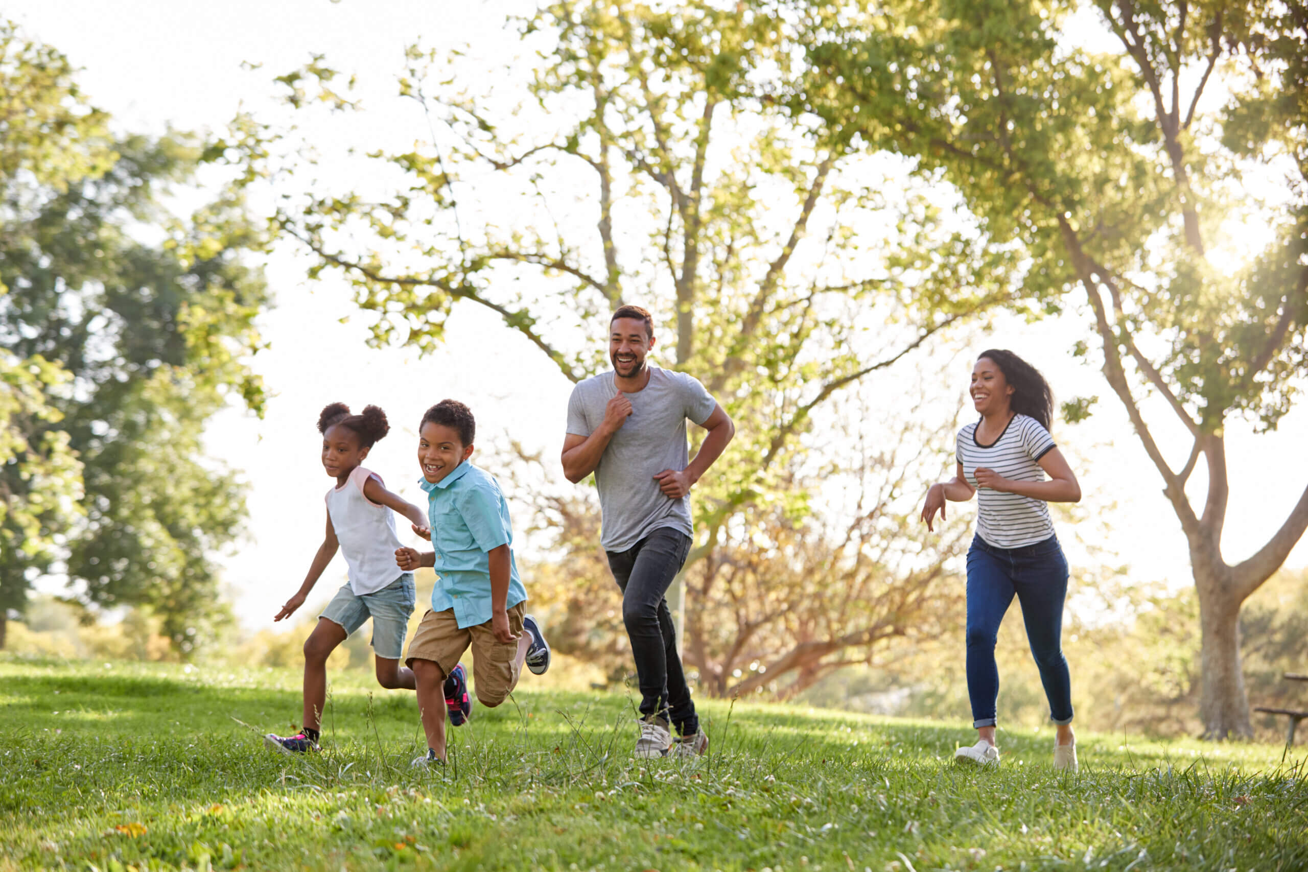 A family, including a mom, dad, boy, and girl, runs and laughs in a park. 