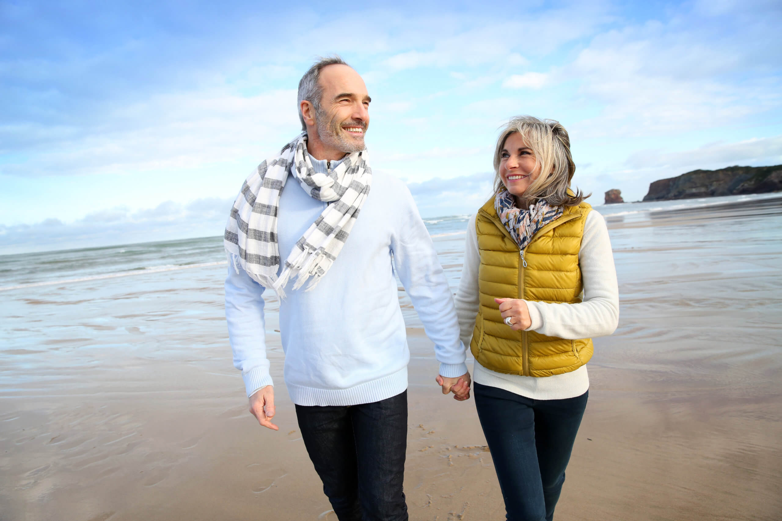 Cheerful older couple walks together holding hands on a beach during cool spring weather.