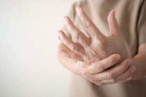 A woman wearing a beige shirt holds her hand in pain due to rheumatoid arthritis symptoms of pain and swelling.