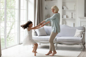 Mature Asian grandmother twirls her young granddaughter around in the living room next to couch.