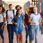 Group of diverse adolescents walks through a school hallway carrying backpacks and smiling.