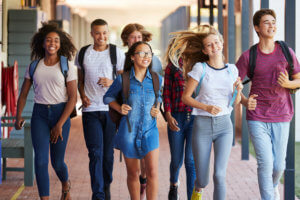 Group of diverse adolescents walks through a school hallway carrying backpacks and smiling.