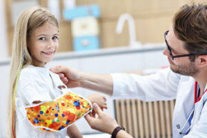 Young blonde girl smiles while a pediatric orthopedic doctor treats her arm with a colorful sling.