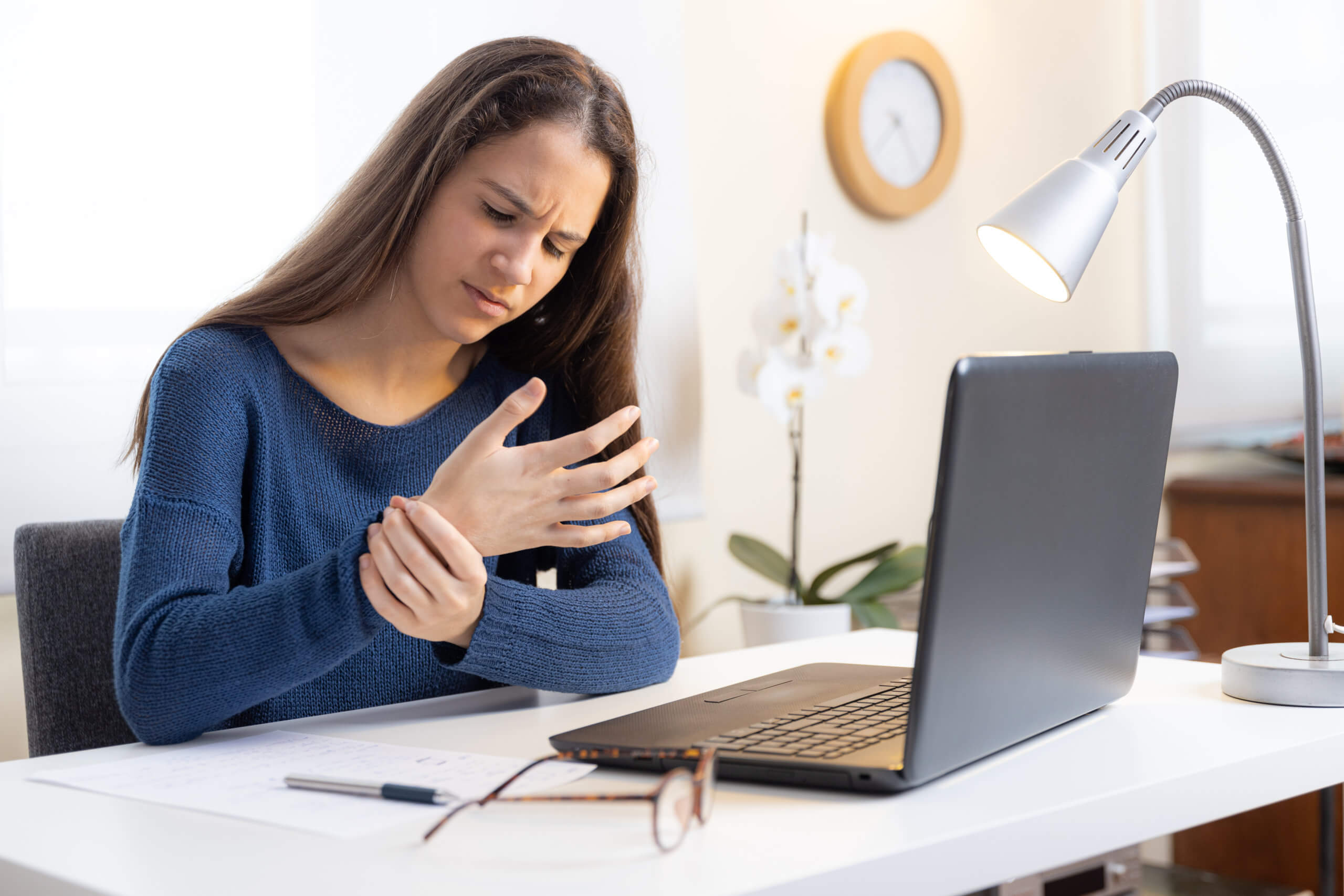 Teen girl working on a laptop holds her wrist and looks in pain due to juvenile arthritis.