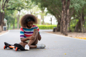 Teen Black girl holds her knee and looks concerned after falling off her skateboard in a park.