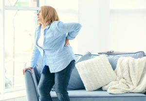 Middle-aged woman holds her lower back in pain while standing and holding on to a sofa.