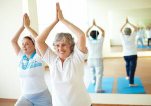 Two older women smile while doing yoga in a sunny studio to stretch their backs.