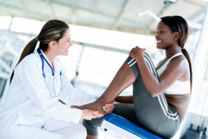 Female orthopedic doctor addresses the ankle injury of a smiling Black athlete wearing sports gear.
