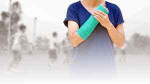 Teen athlete in blue shirt holds her green cast with her free hand as sports players play a game in background.