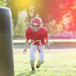 Young child in red-and-white football uniform and helmet runs on the green grass playing field toward tackling dummy.