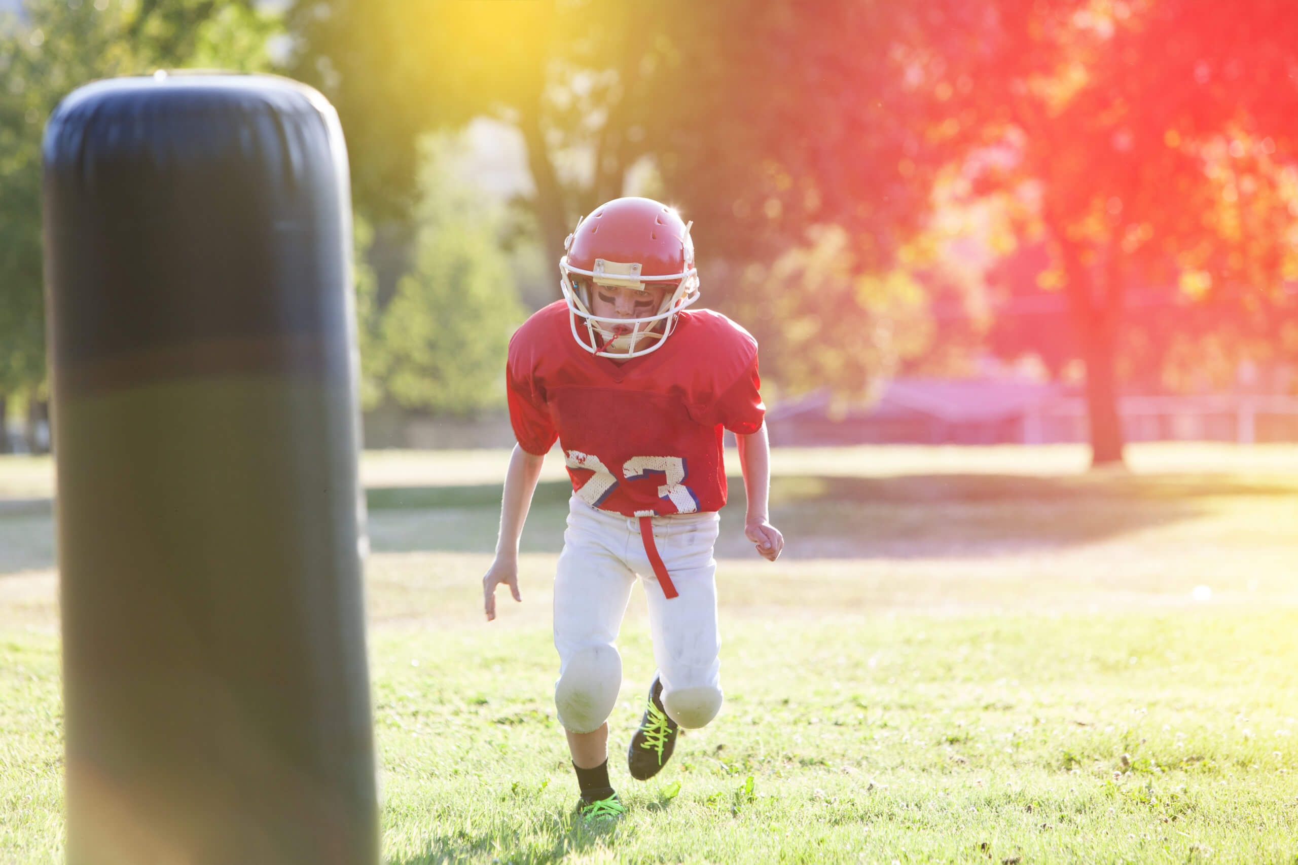 Young child in red-and-white football uniform and helmet runs on the green grass playing field toward tackling dummy.