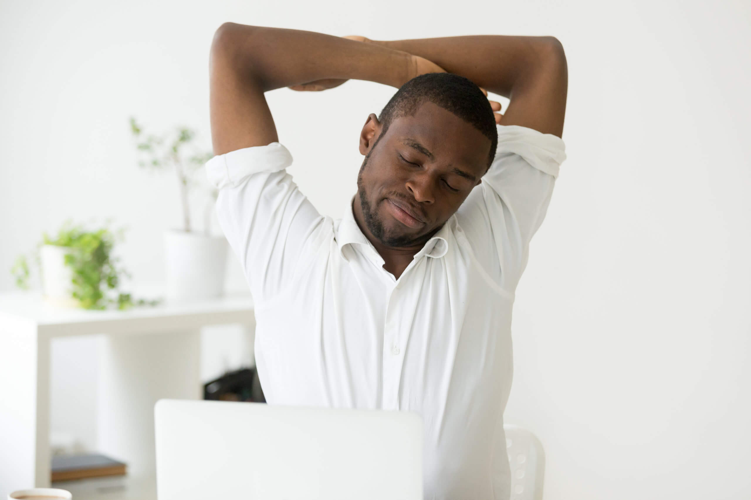 A young Black man stretches his arms while at his desk to relieve upper body pain.