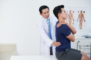 A young man performs an upper body posture exercise while a doctor examines his back.