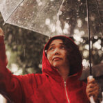Woman in red coat walks in a park with an umbrella during a rainstorm.