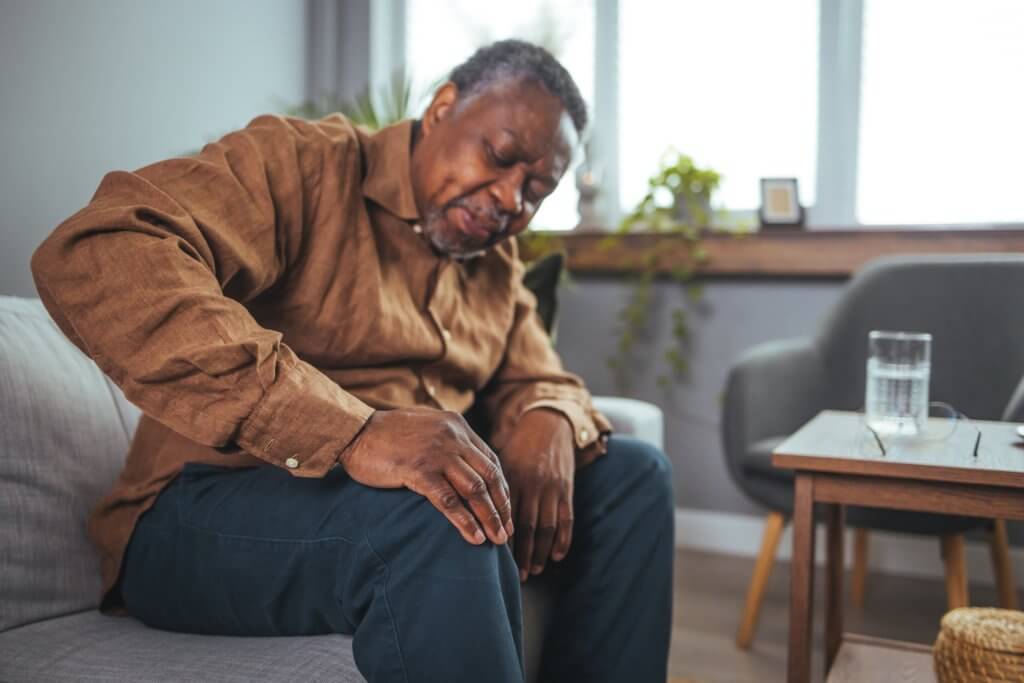 Older man holds his knee while sitting on the couch at home. 