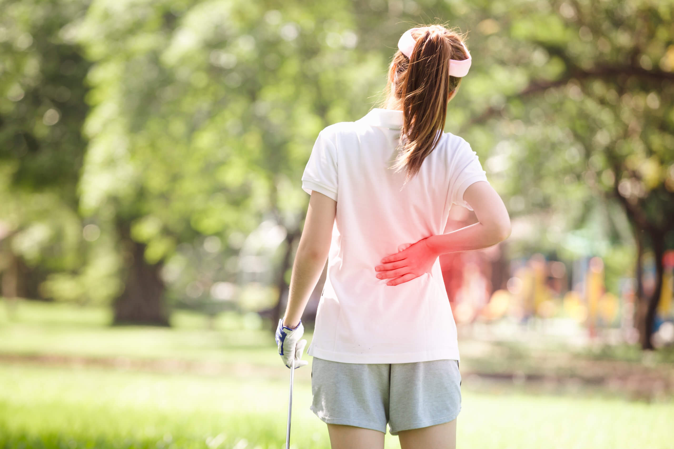 A woman, with her back to the camera, holds a golf club in one hand and her lower back with the other.