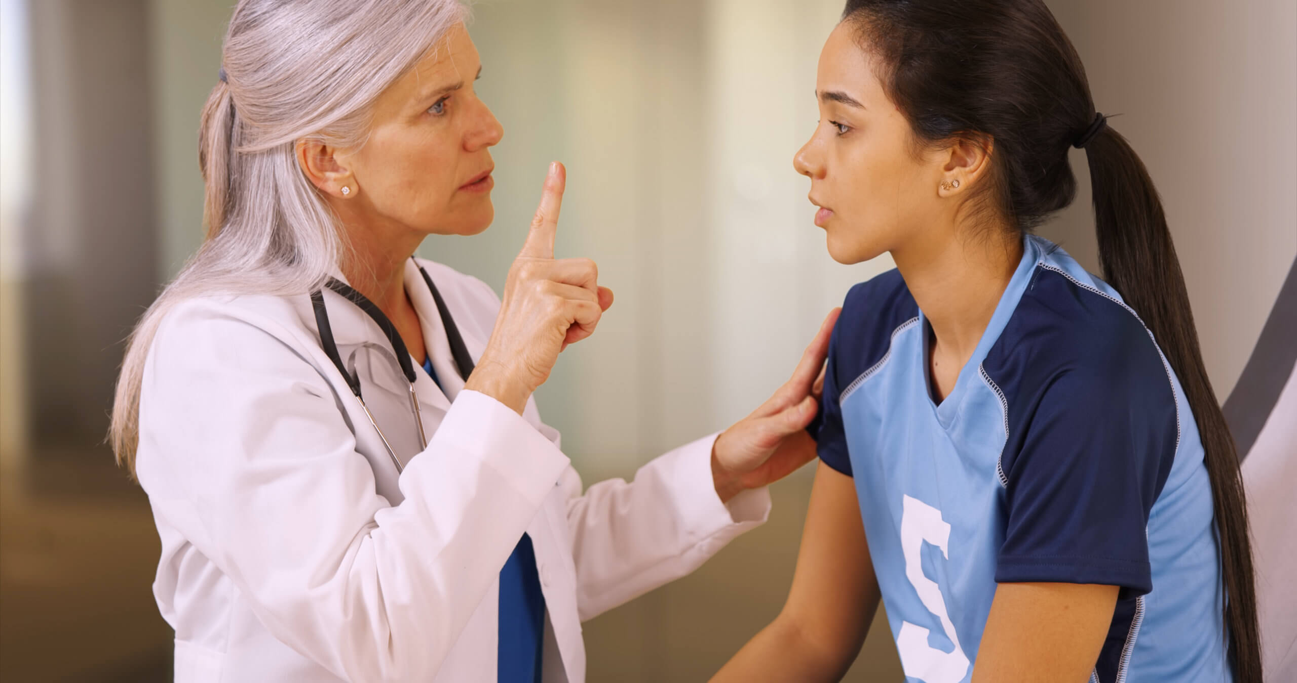 A doctor stands in front of a teen girl in a soccer jersey, holding up one finger and checking for signs of a concussion.