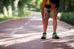 A runner stops in the middle of a path and holds their leg and knee in pain.
