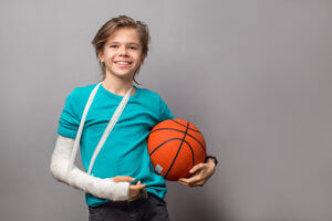 A child holds a basketball in one arm while the other arm is in a cast and sling.