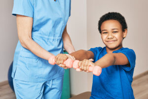 A young boy holds up two weights as a physical therapist assists him.