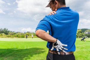 A man stands on a golf course and holds his lower back and shoulder in pain.