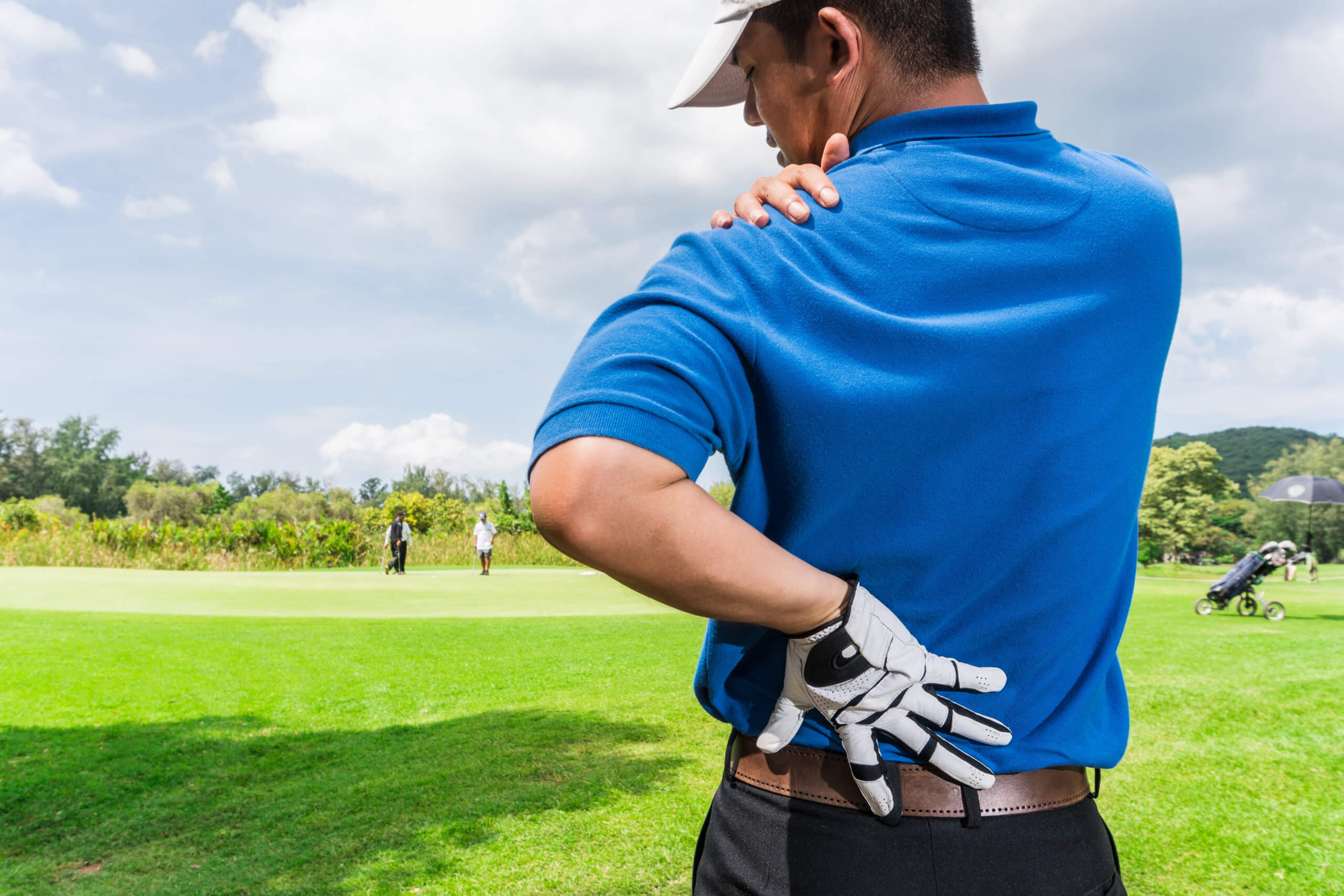 A man stands on a golf course and holds his lower back and shoulder in pain. 