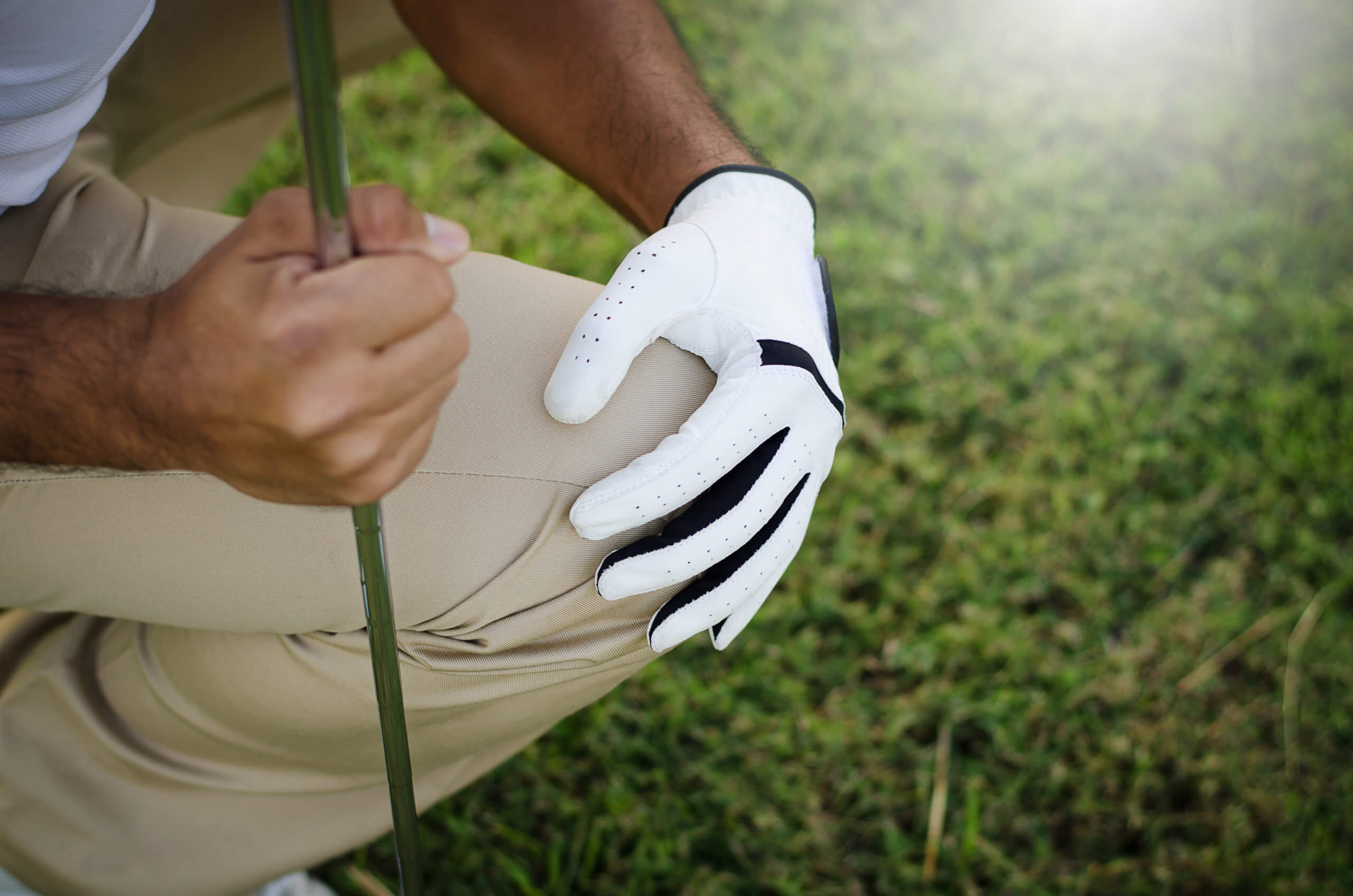  A male golfer crouches down in the grass, holding a club with one hand and his knee with the other. 