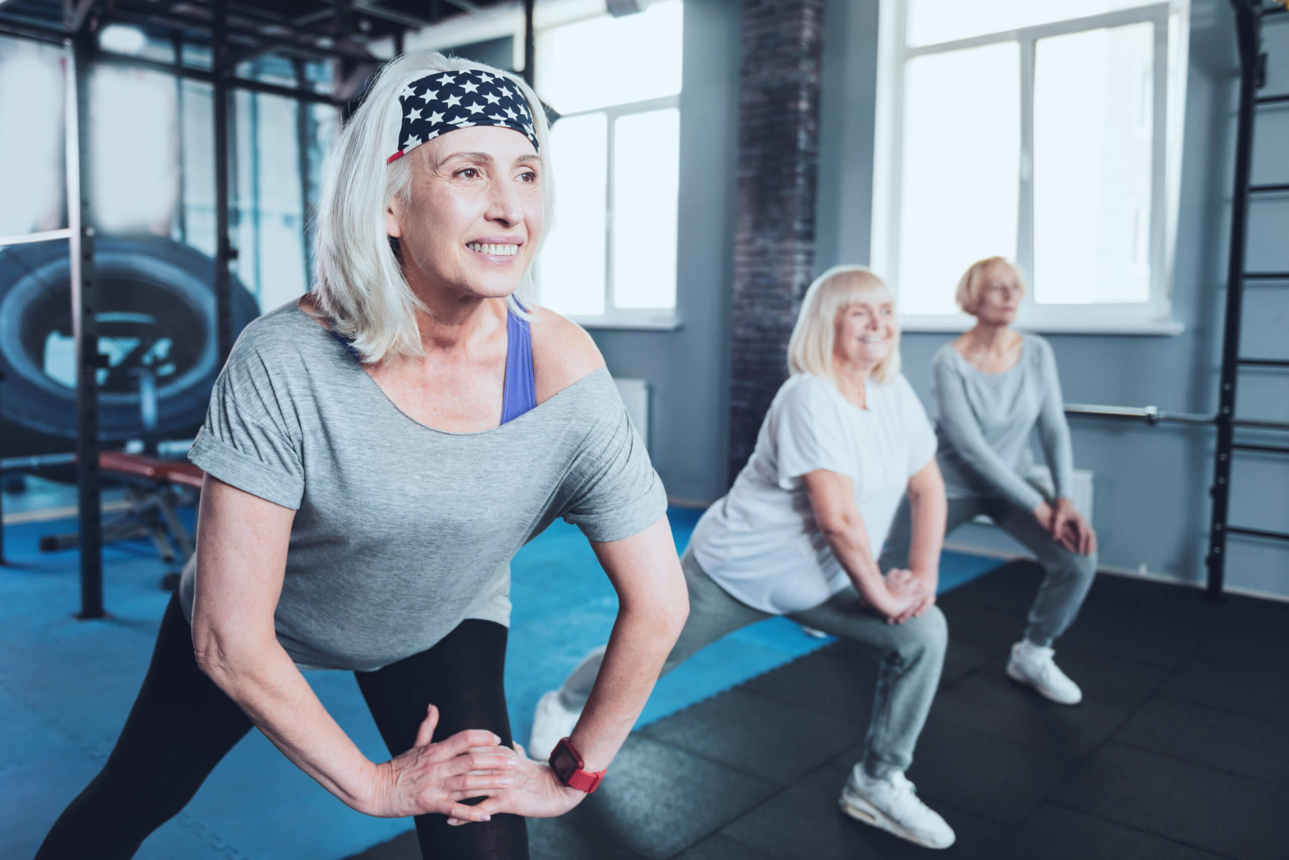 Three older women in athletic clothing do lunges as part of a stretching routine in a gym.