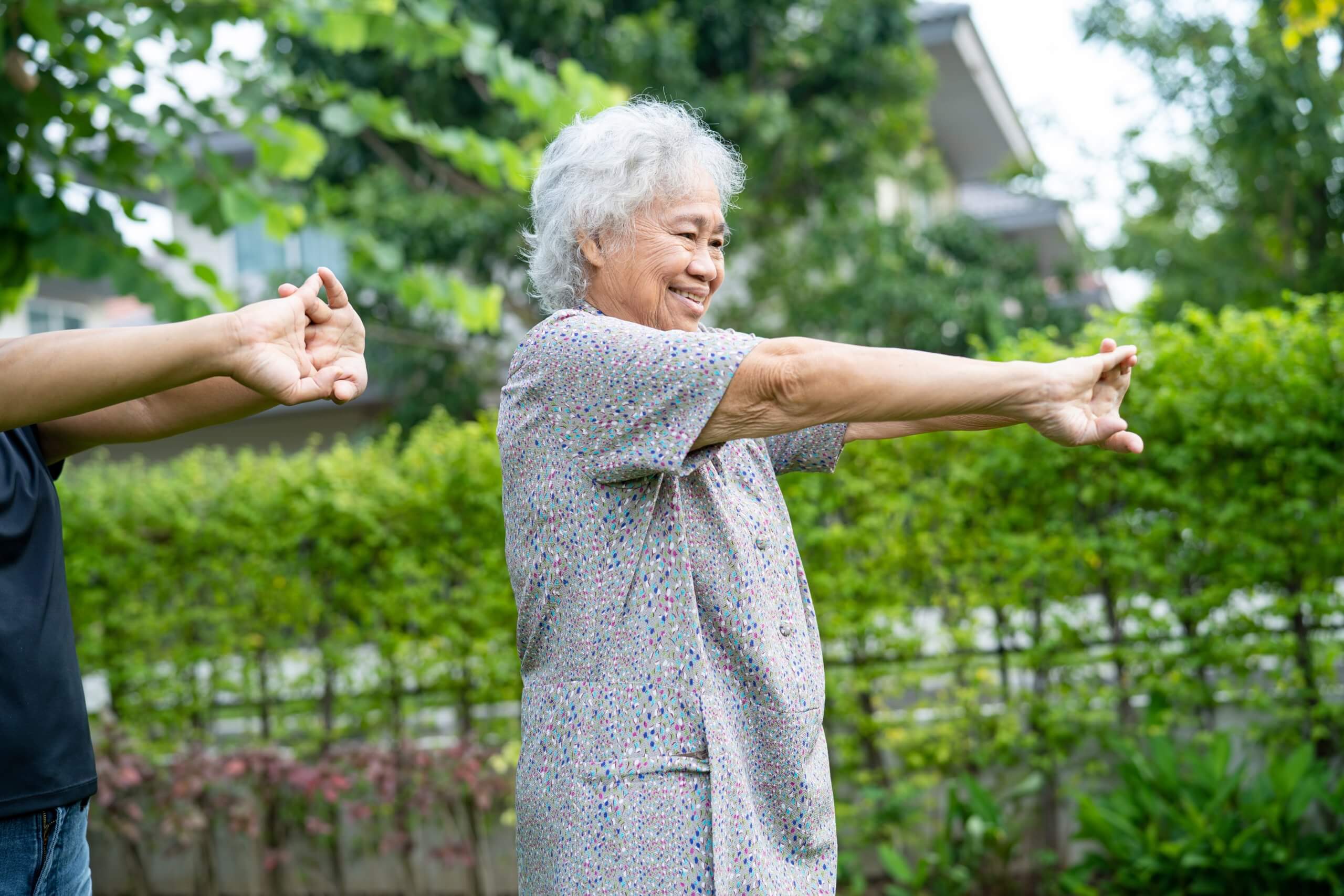 An older woman stretches her shoulders and arms in a park. 