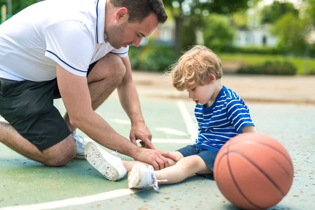 A dad assesses his young son’s leg as they sit on a basketball court. 