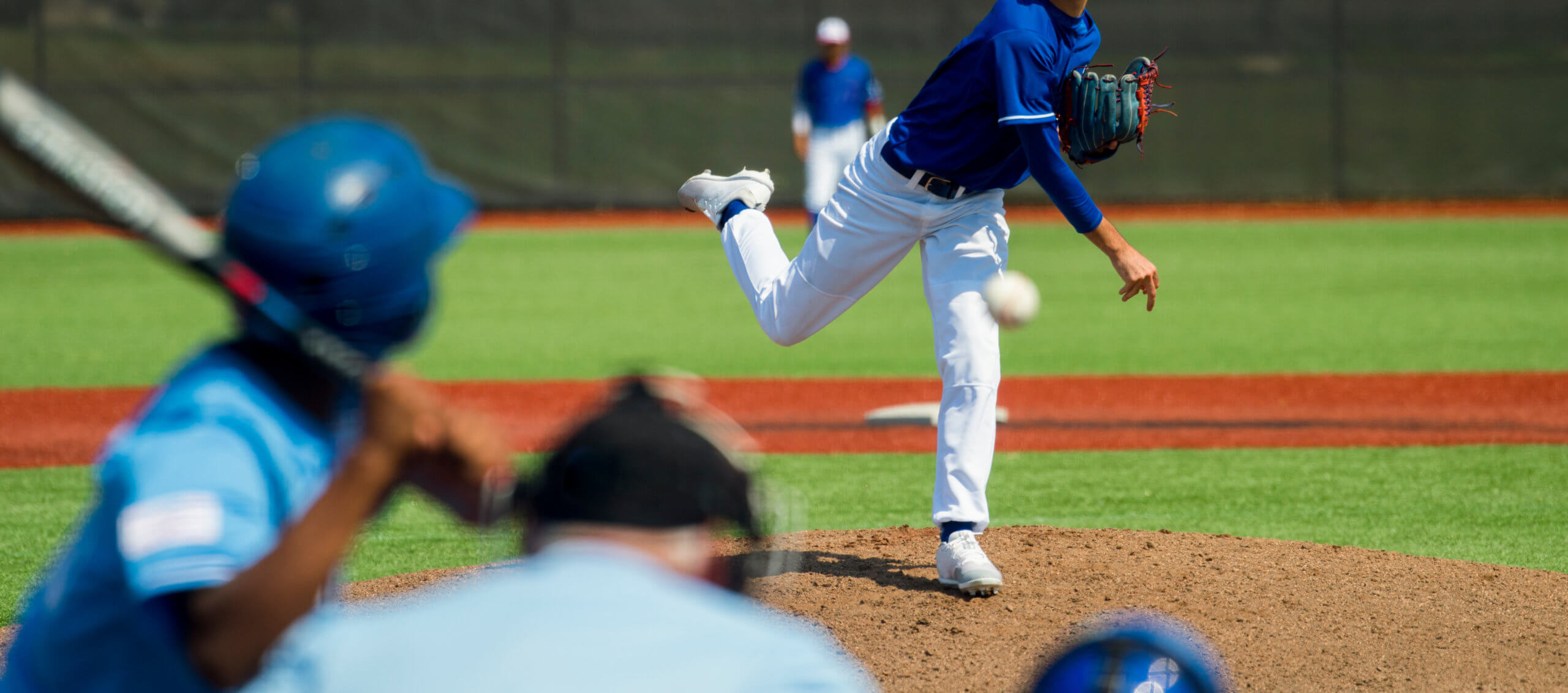 Pitcher and third baseman in blue jerseys during a baseball game. Pitcher has thrown the ball to the batter. The pitcher, batter, catcher, and outfield are all visible in the picture.