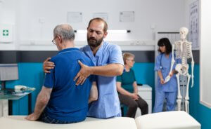 A male medical professional examines an older man’s back as he sits on a table.