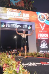 Dr. Garrett Moss crossing the finish line of the IronMan World Championship. His name, number: 2158, and time - 10:46:04, is displayed on the scoreboard behind him.