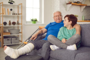 A boy sits on the couch next to an older man with his casted leg and foot elevated.