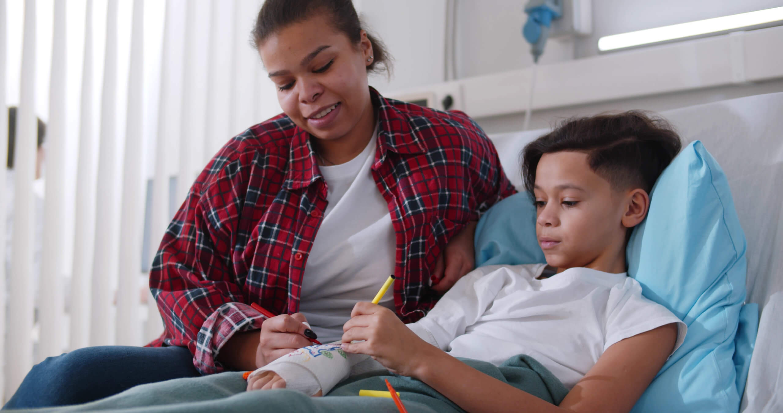 A woman sits next to a boy in a hospital bed as they draw on his cast.