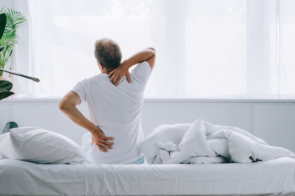  Man sitting on bed holding his lower back in pain as he wakes up. 