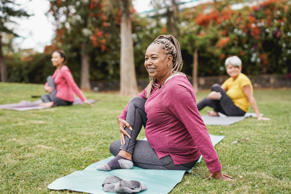 An older woman stretches and smiles on a yoga mat in a park. 