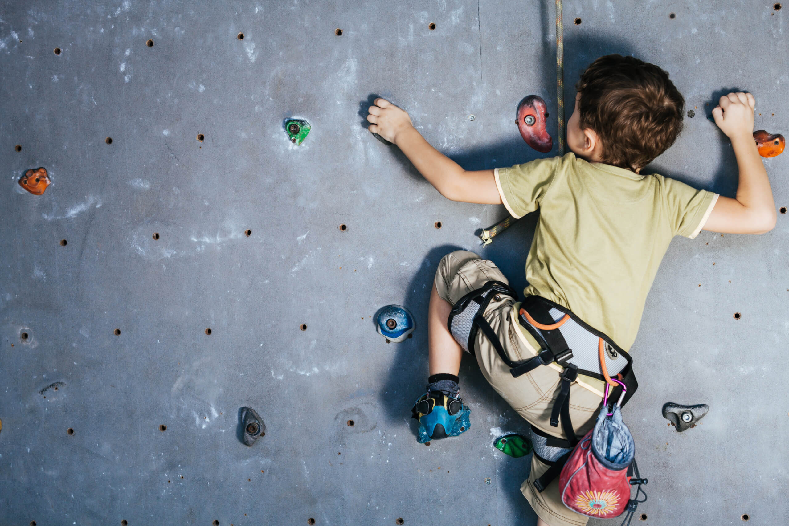 A young boy climbs a climbing wall wearing a harness and chalk bag. 