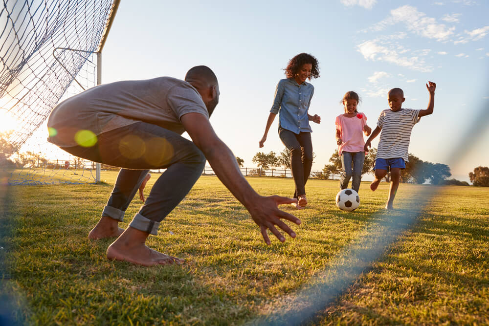A mother, father, son, and daughter play soccer. The girl kicks the ball toward the dad in the goal. 