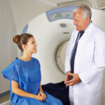A doctor prepping his patient as she sits on the MRI machine bed.