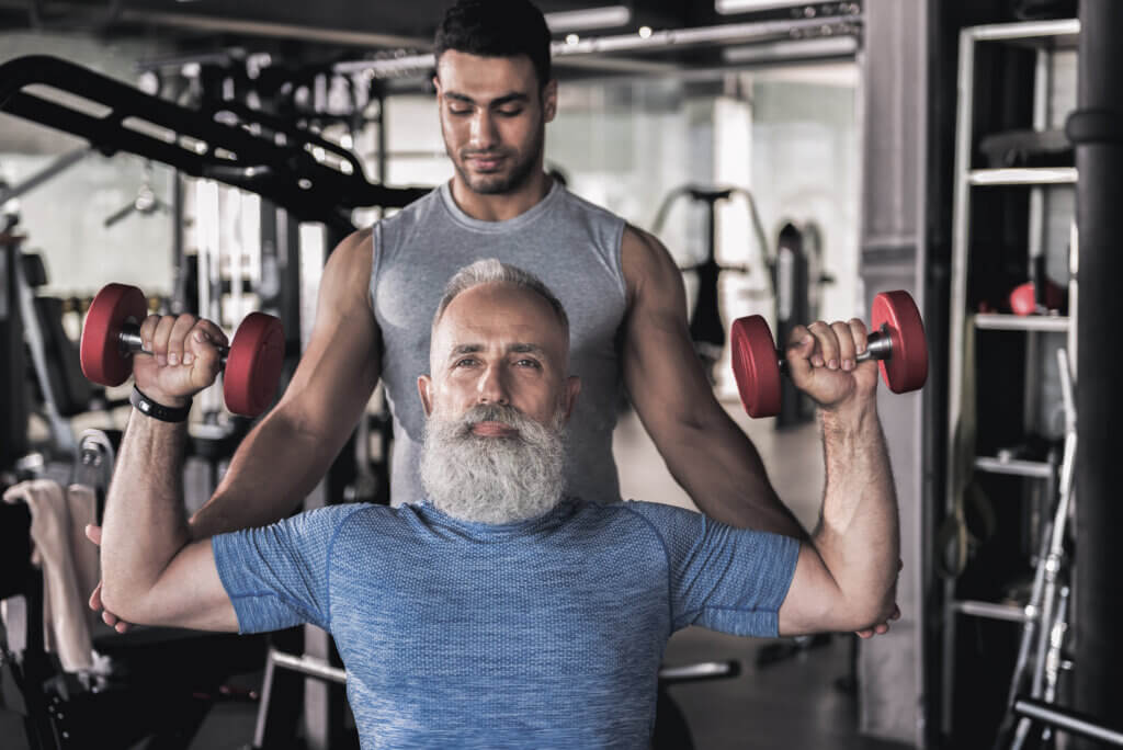 Photo of stylish old bearded man is sitting in modern gym and raising dumbbells over his head. His personal trainer is touching his elbows while standing behind him, guiding him through the workout. Senior sportsman doing exercises in athletic center with his instructor