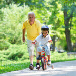 Happy grandfather playing in the park with his grandson. Grandson is on his bike and grandfather is running behind him