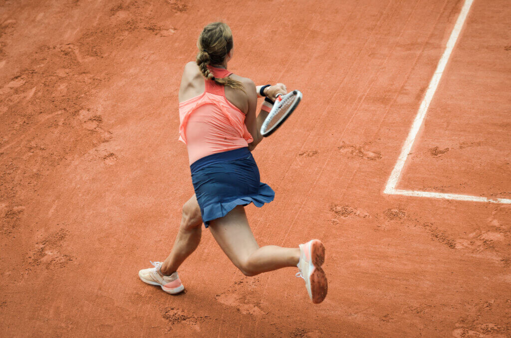 Back view of a woman playing backhand in tennis outdoors on a clay court. Woman has recovered from a quadriceps tendon injury