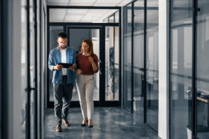 Two formally dressed employees taking a stroll down an office hallway while looking at an Ipad.