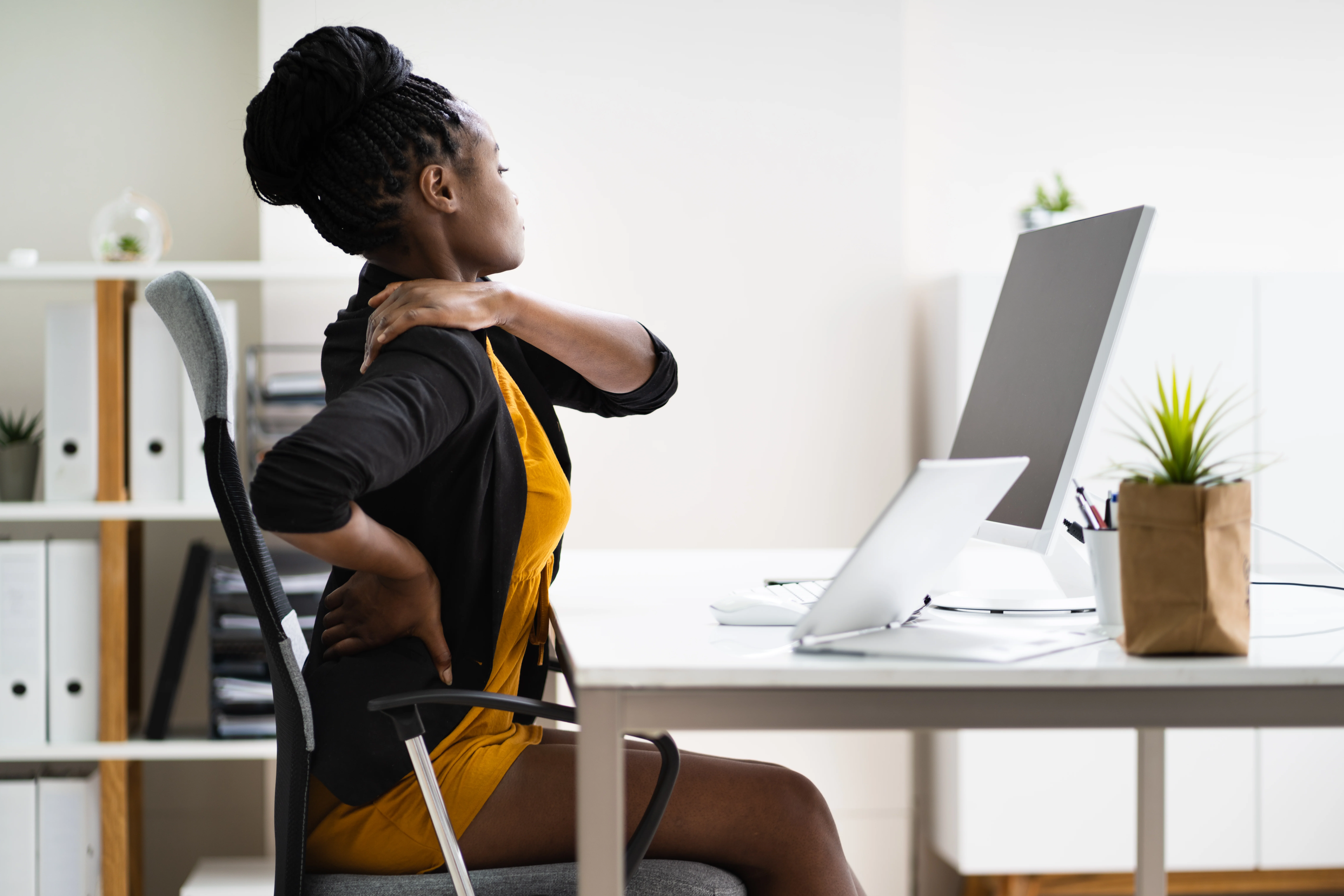 Professional Black woman stretches and massages her back while sitting at a desk working on a computer. 