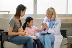 A young girl in an arm cast sits on a couch next to her mother and a female doctor.
